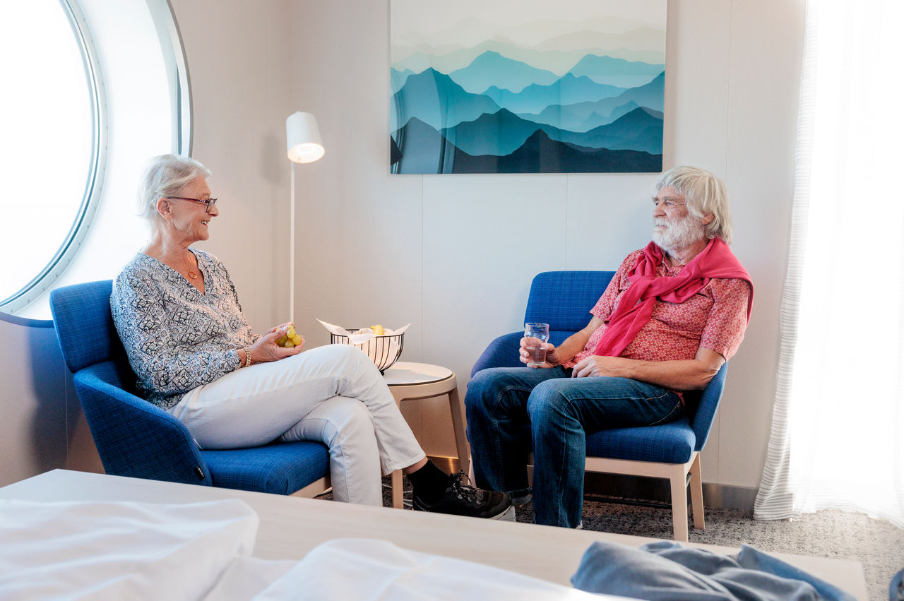 An older couple sitting facing each other, the man holding a drink, the woman some green grapes, relax in a cabin.